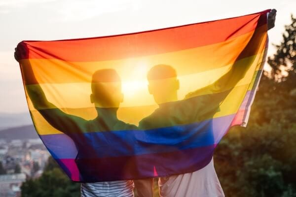 Imagem de um casal do mesmo sexo em frente ao arco-íris, símbolo da comunidade LGBTQIA+